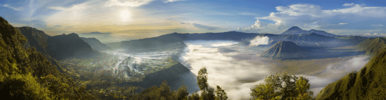 A morning panorama view of Mount Bromo and the surrounding valleys and landscape, showing mountains covered trees and dressed in clouds that seem to stretch on forever